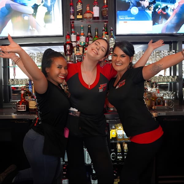Three smiling Jaks Bar & Grill bartenders pose happily behind the bar with arms raised.