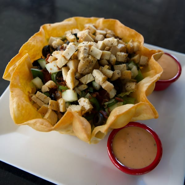 A taco salad in a crispy tortilla bowl with two red ramekins of dressing on a white plate.