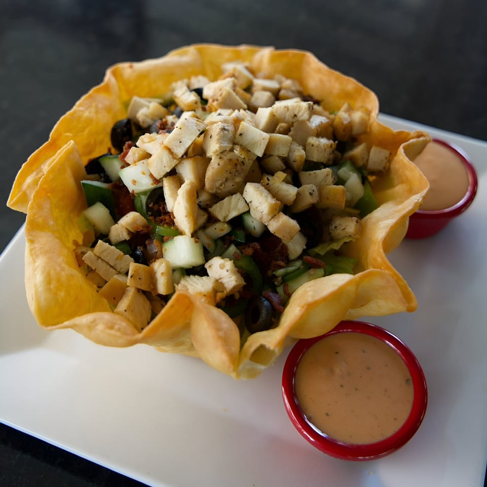 A taco salad in a crispy tortilla bowl with two red ramekins of dressing on a white plate.