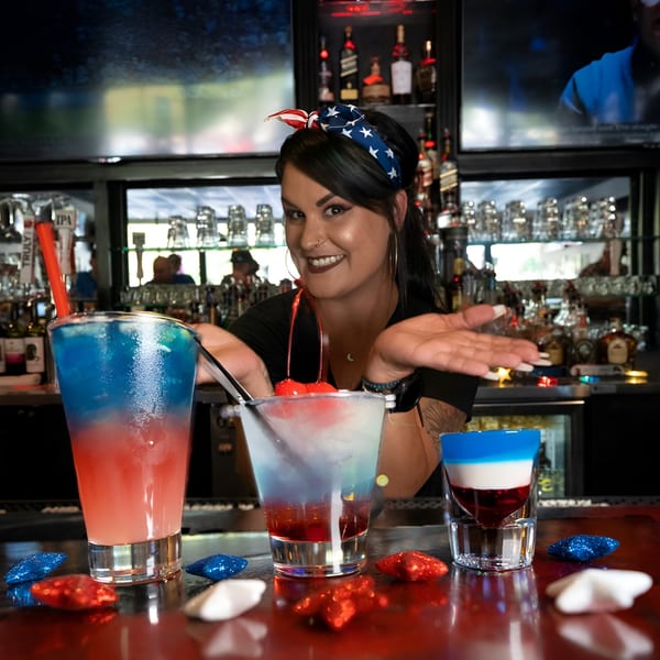 Bartender smiles behind a bar with three red, white, and blue drinks and star decorations.