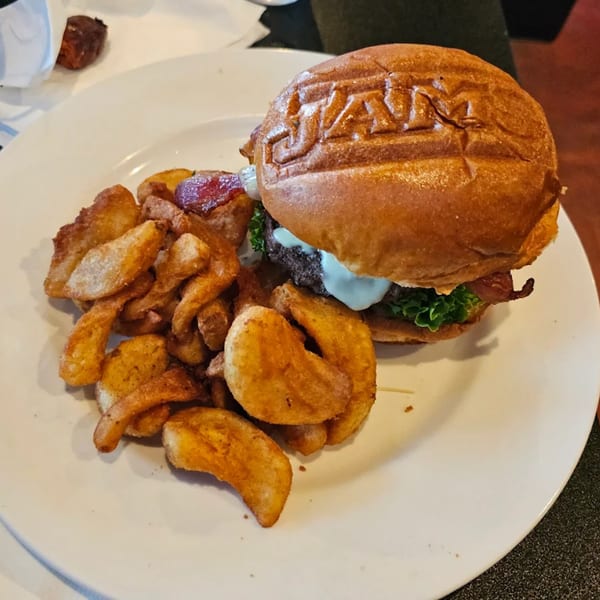 A Jams@209 burger with a branded bun, bacon, lettuce, and sauce, served with seasoned potato wedges.