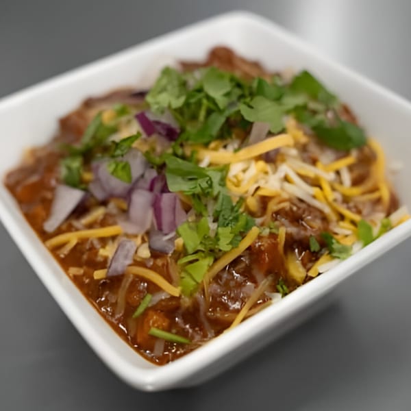 A white bowl of Jams@209 chili topped with shredded cheese, diced red onions, and cilantro.